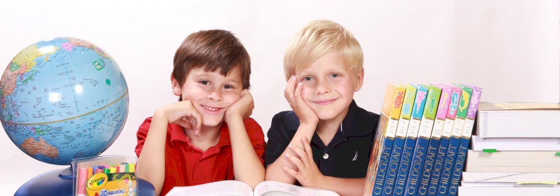 Zwei Jungen sitzen an einem Tisch inmitten von Büchern, Schreibutensilien und einem Globus.